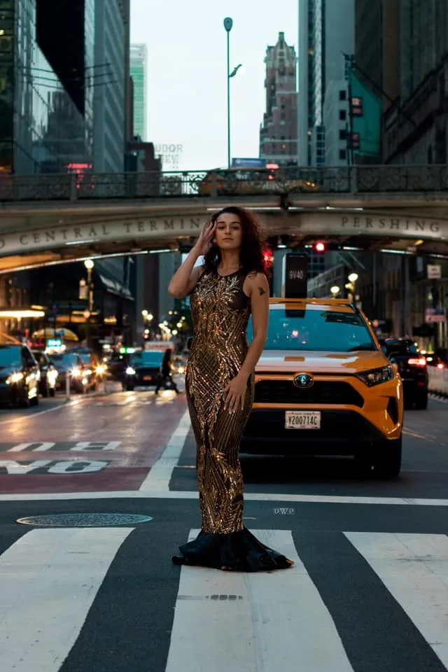 Model in a gold patterned gown standing in a crosswalk, a taxi behind.